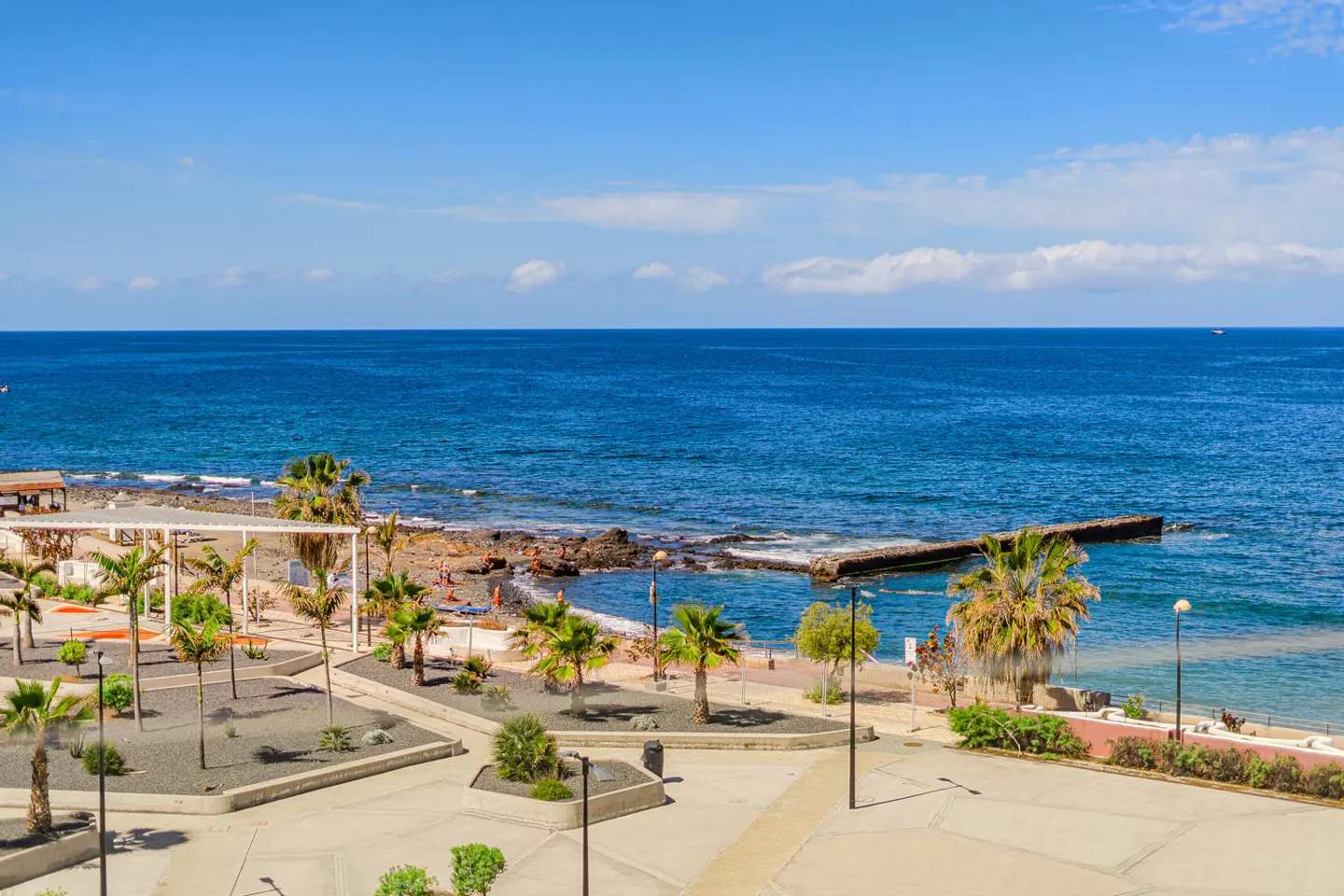Ocean view with palm trees, beach, and blue sky. A concrete walkway leads to the beach.