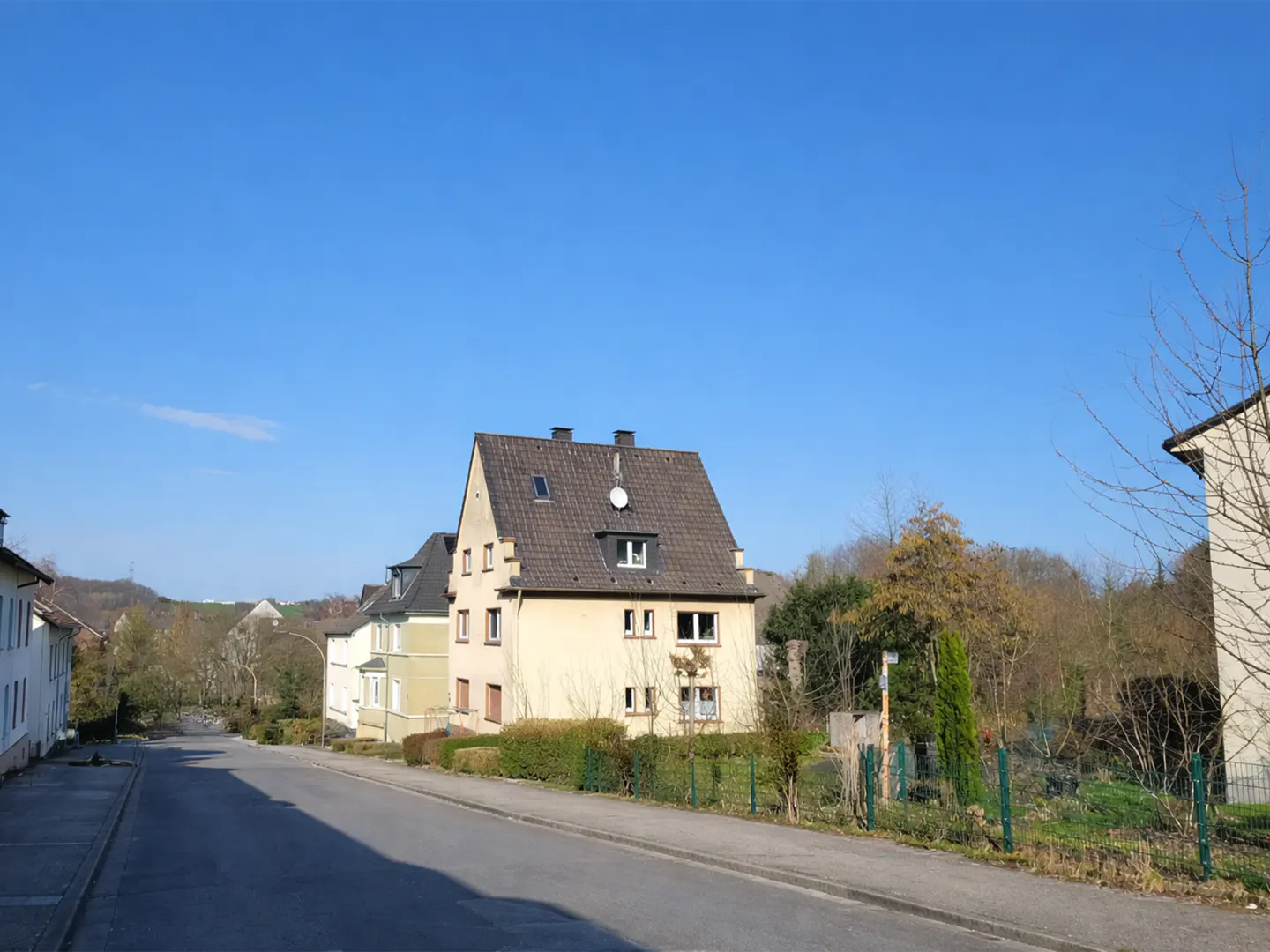 Street view of a light yellow two-story house with a brown roof and a satellite dish under a clear blue sky.