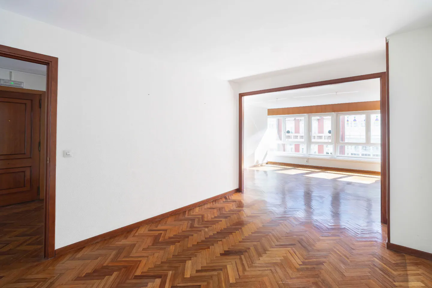 Empty room with white walls, herringbone wood floor, and a doorway to a bright room with large windows.