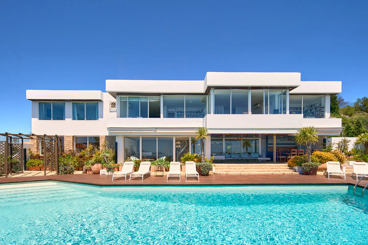 Modern white house with a pool. The house has large windows and a wooden deck with lounge chairs. Blue sky above.