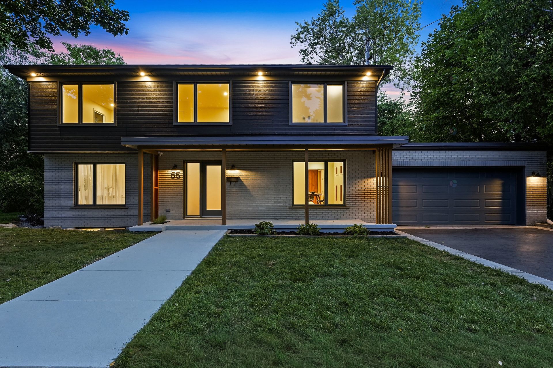 Two-story modern home with black upper siding, grey brick lower level, attached garage, and illuminated windows at dusk.