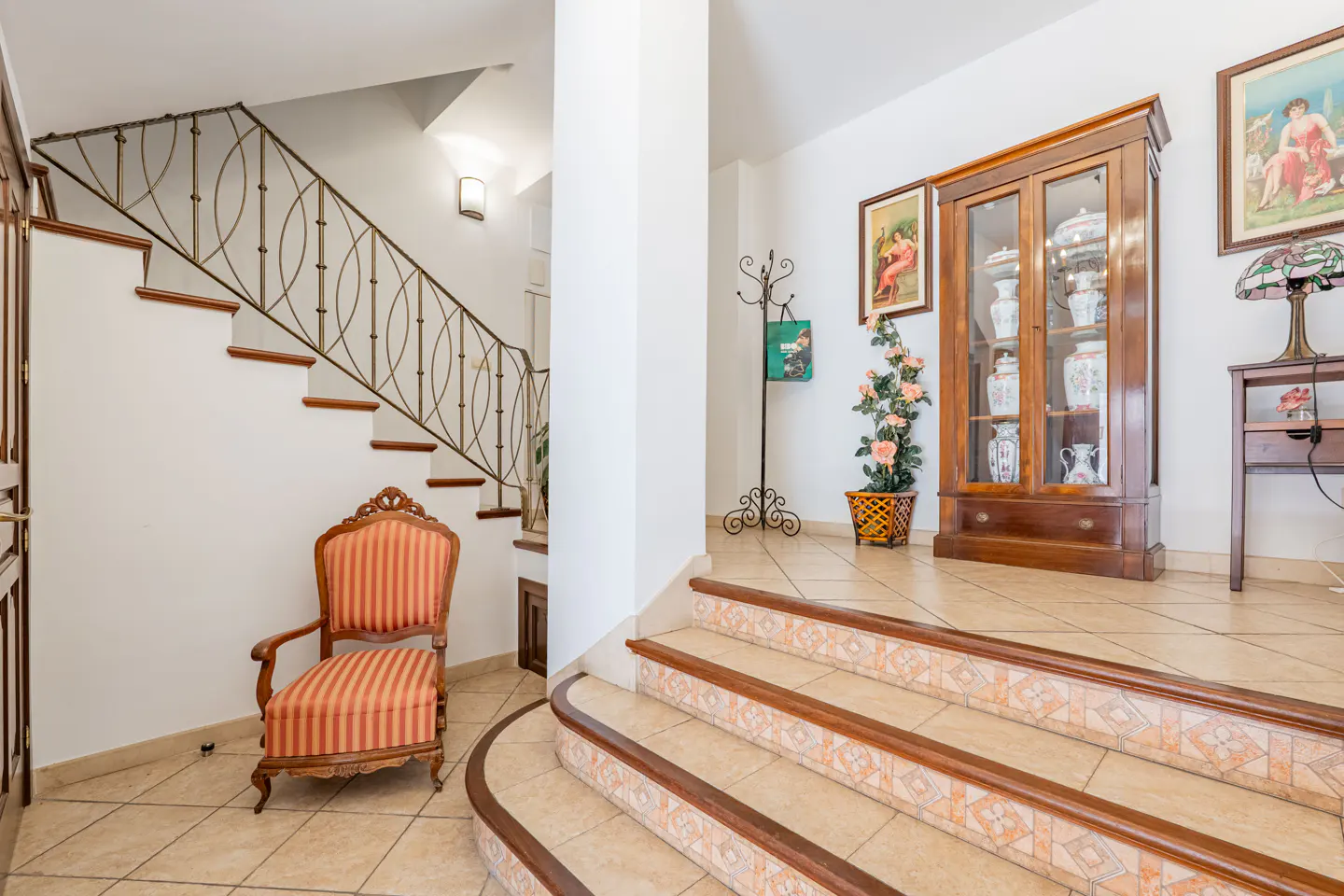 Foyer with tiled stairs, a striped armchair, a wooden display cabinet with vases, and a staircase with a metal railing.