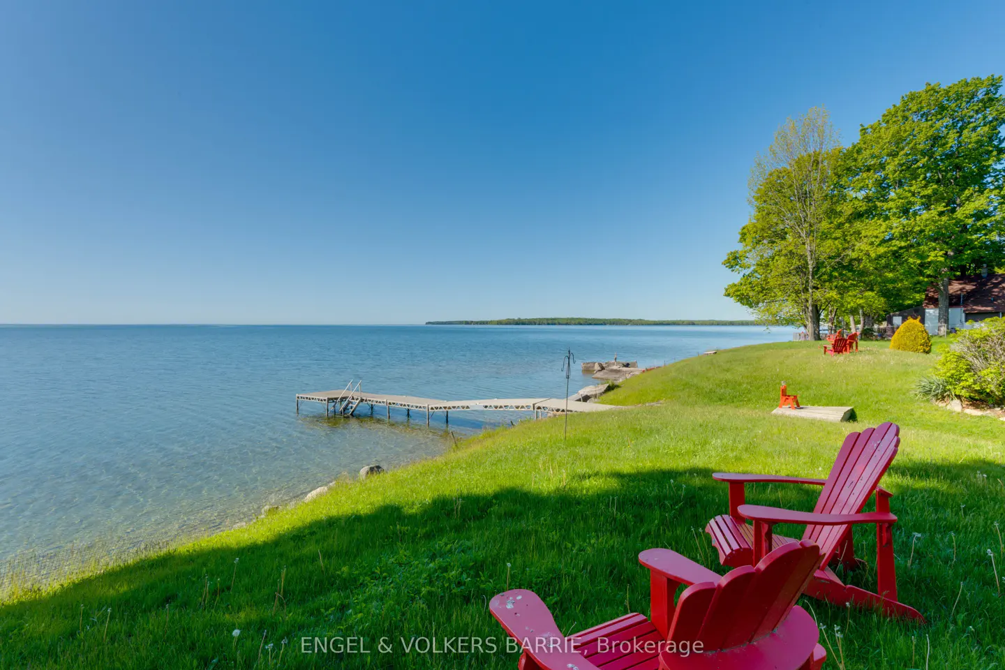 Waterfront property with two red Adirondack chairs on green grass overlooking a lake with a dock under a clear blue sky.