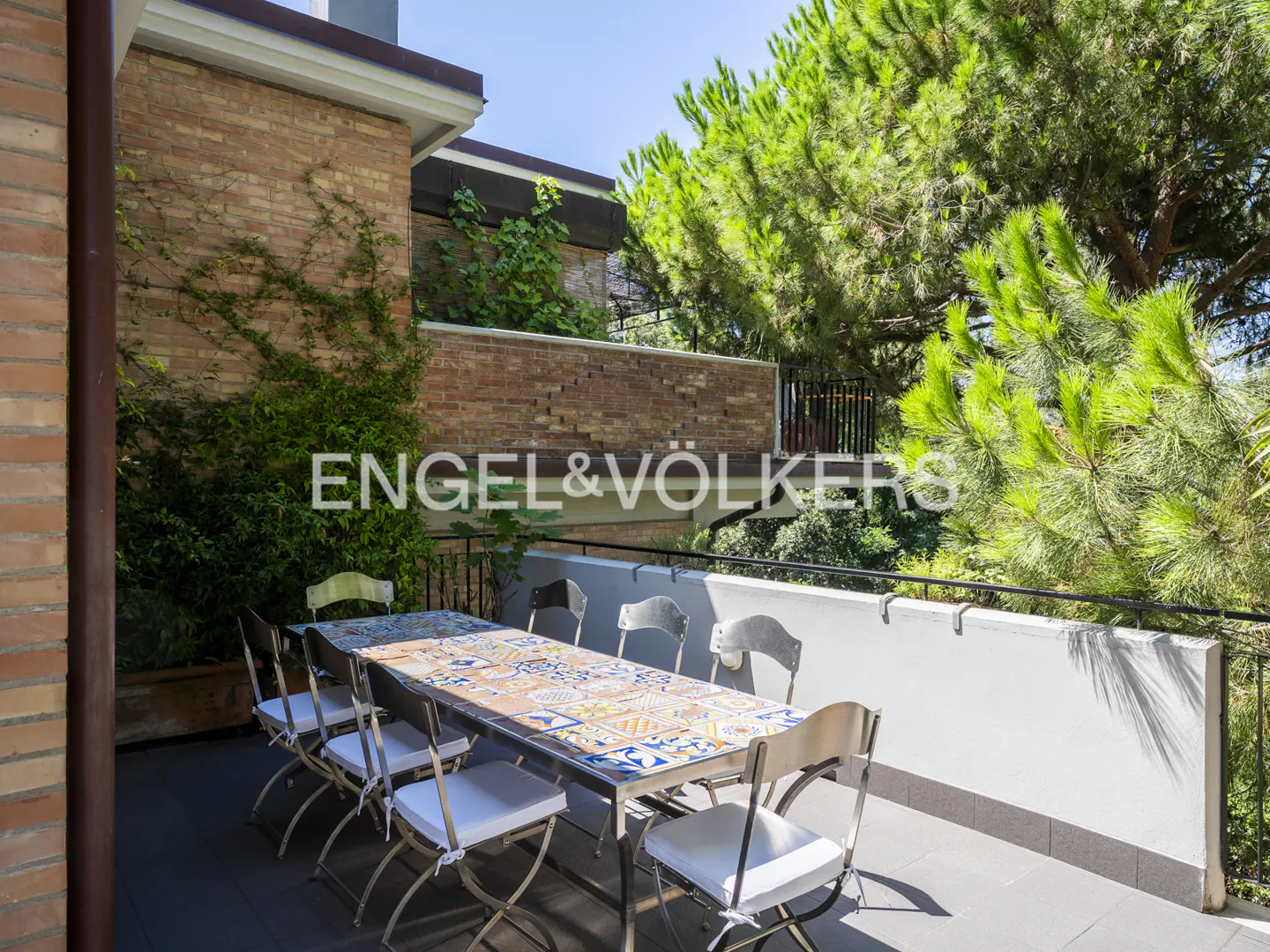 Outdoor patio with a long table, chairs, and a brick building with green plants in the background.