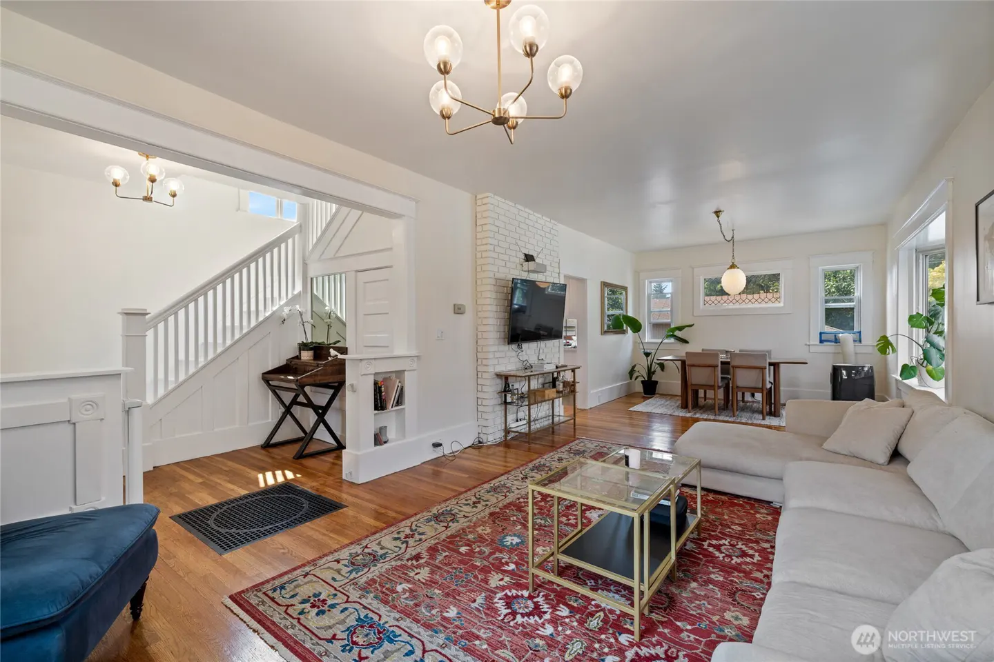 Bright living room with hardwood floors, a red patterned rug, and a white sectional sofa. A staircase is visible in the background.