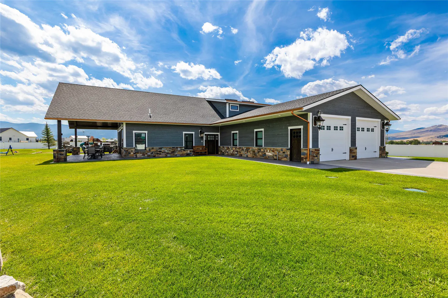 Modern gray home with stone accents, two-car garage, and covered patio, set against a large lawn under a blue sky.