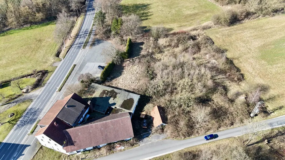 Aerial view of a property with a red-roofed house, adjacent lot, and road. A blue car drives on the road.