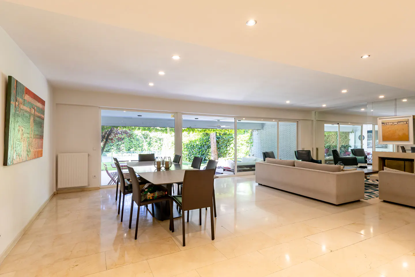 Bright, open-plan living and dining area with marble floors. A dining table with chairs sits near a large window overlooking a green garden. A beige sofa is in the living area.