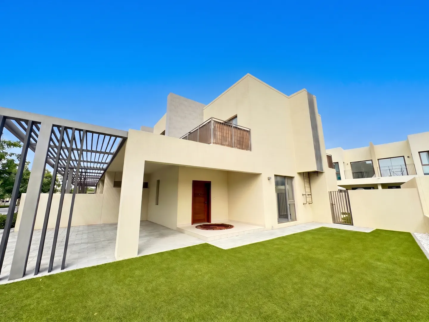 Modern two-story beige house with a brown door, a small balcony, and a green lawn under a clear blue sky.