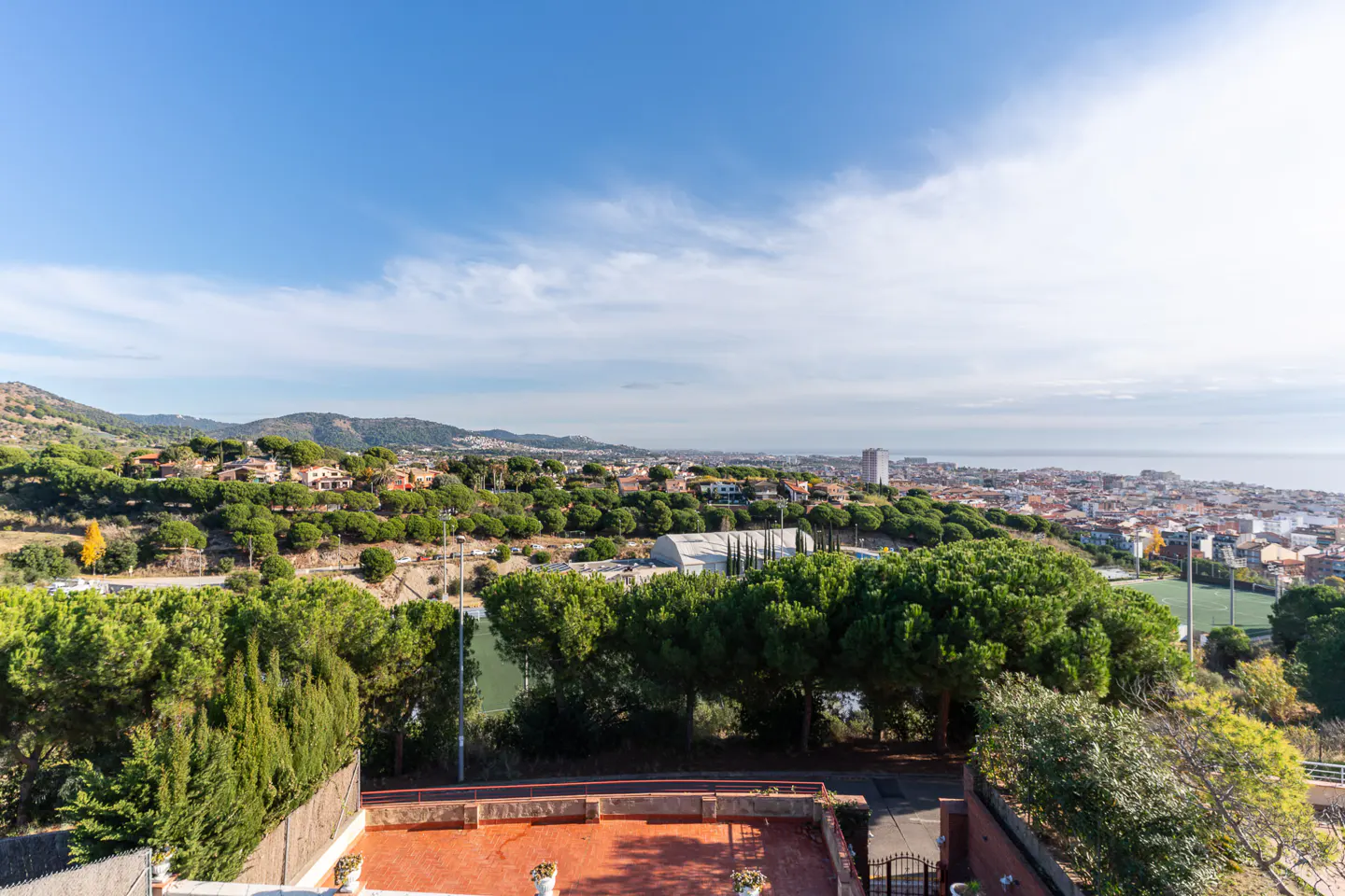 View from a red-tiled patio overlooking a green landscape with trees, a city, and the ocean under a blue sky.