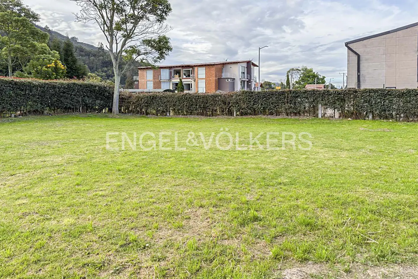 A green lawn with a hedge in the background, a tree on the left, and a building in the distance. The Engel & Völkers logo is superimposed on the lawn.