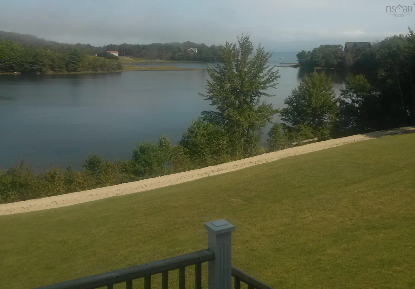 Water view from a grassy lawn with a wooden railing in the foreground. Trees line the shore.