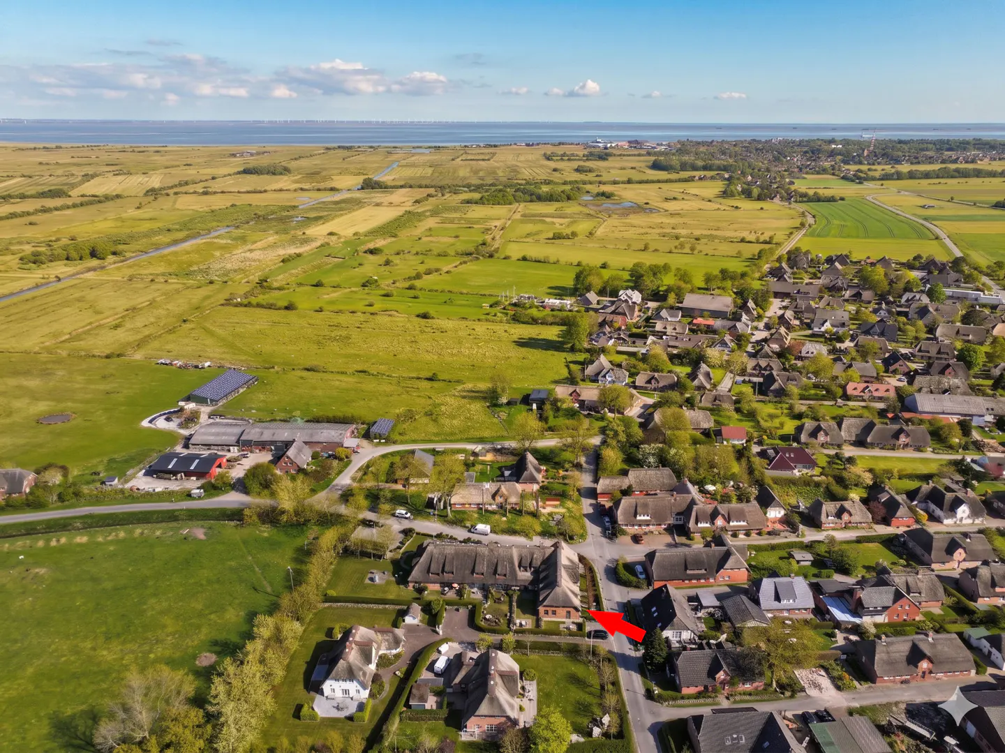 Aerial view of a village with thatched-roof houses, green fields, and the sea in the background under a blue sky.