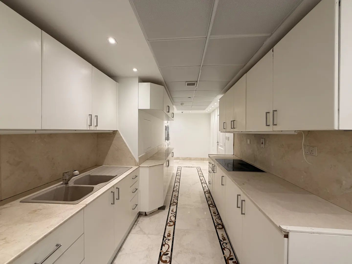 A long, narrow kitchen with white cabinets, marble countertops, and a decorative tile floor.