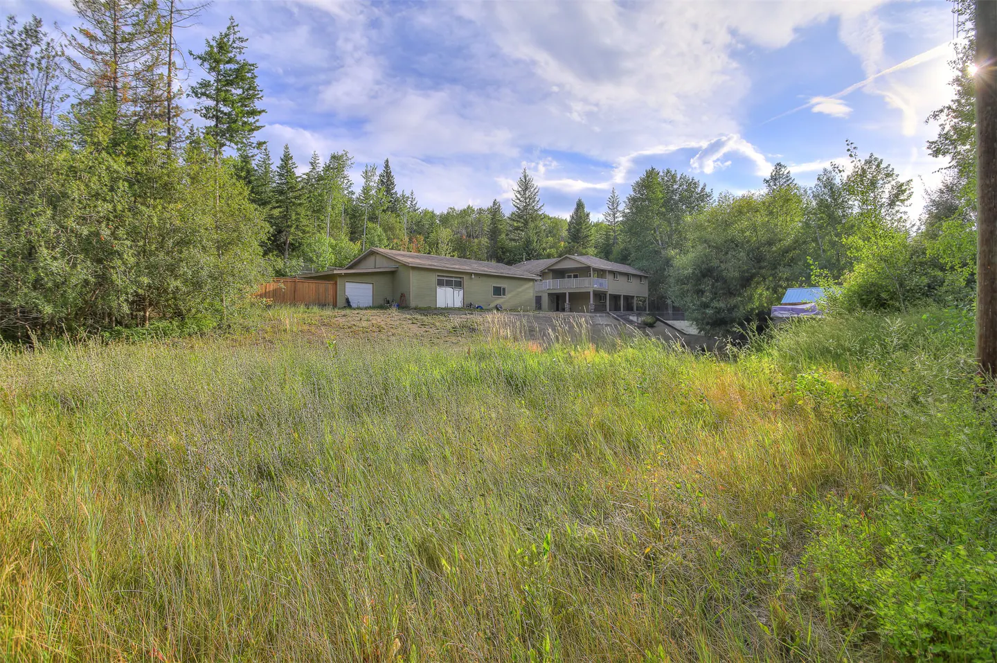 A two-story house with a garage is surrounded by tall grass and trees. The sky is blue with white clouds.