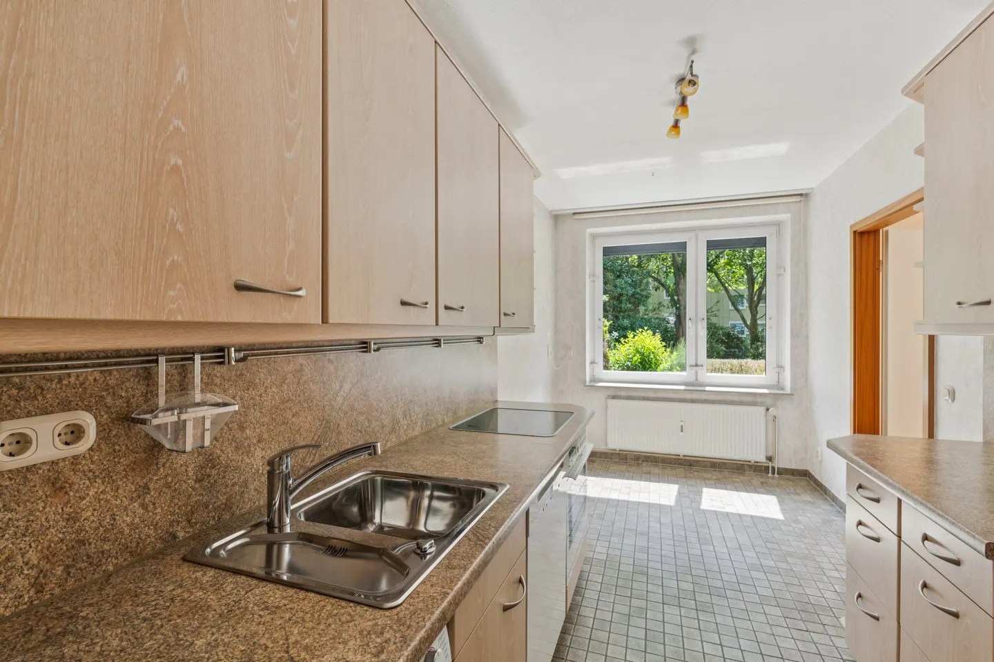 A kitchen with light wood cabinets, a granite countertop, and a stainless steel sink. A window overlooks a green yard.