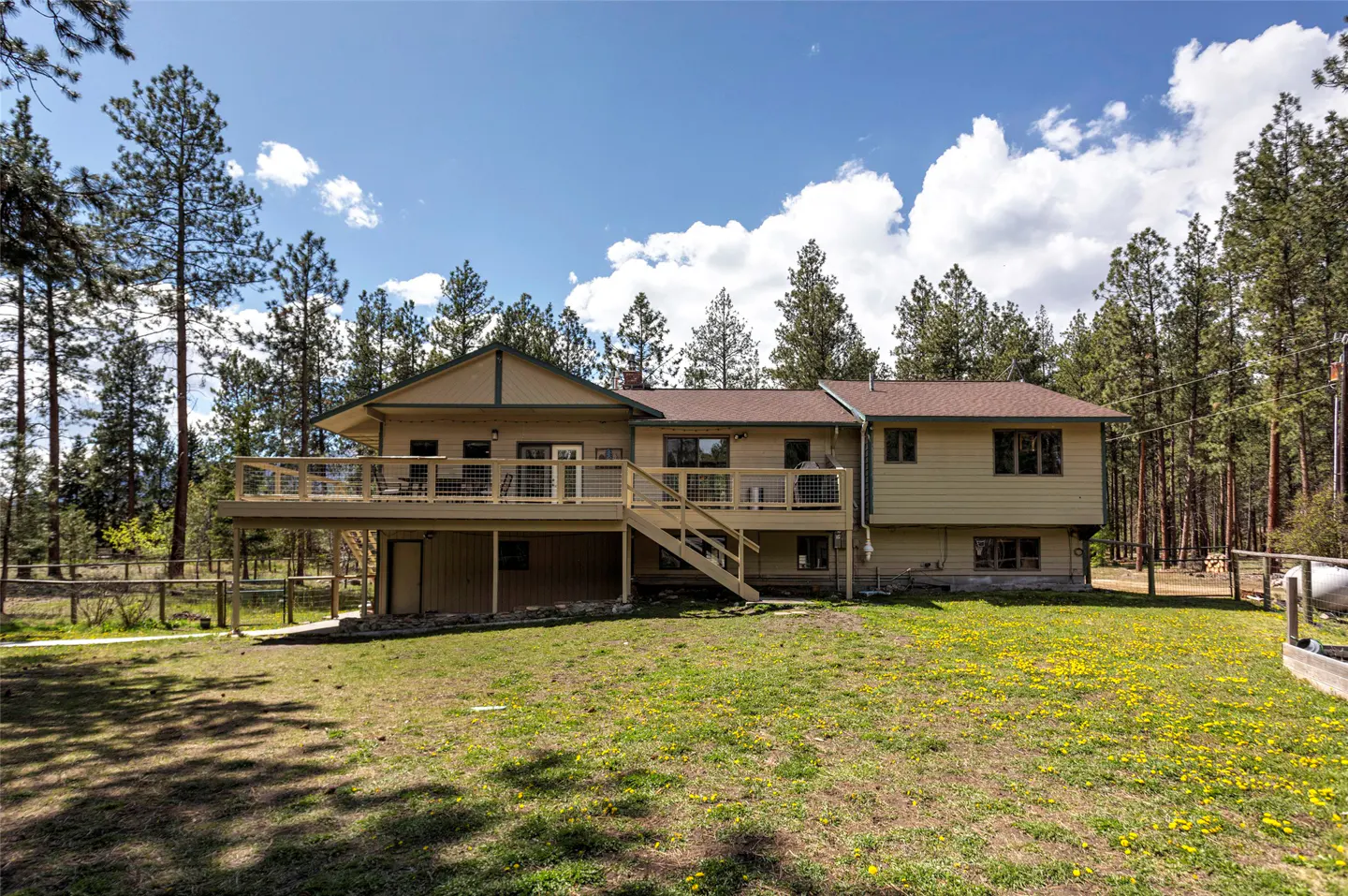 Tan house with a brown roof, a wooden deck, and stairs, surrounded by a green lawn and tall trees under a blue sky.