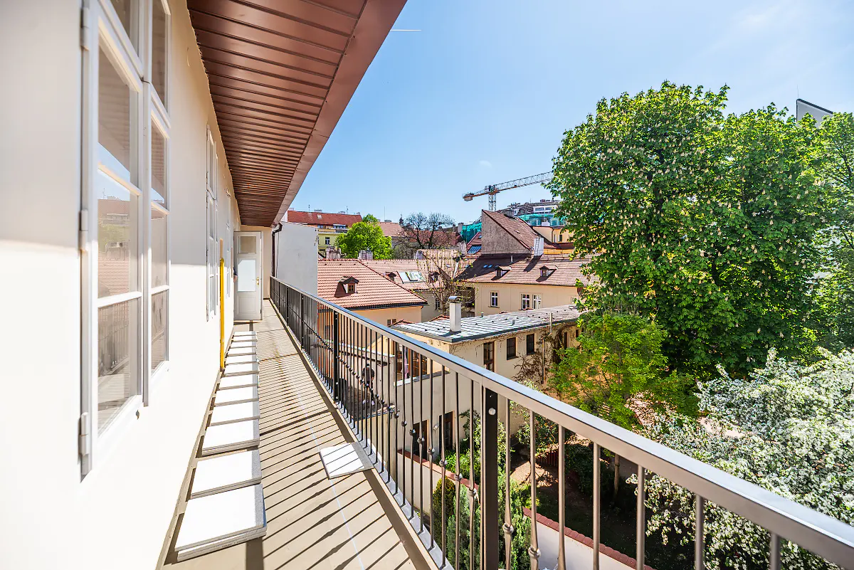 Balcony view with metal railing, white building, and city rooftops. Green trees and blue sky in the background.