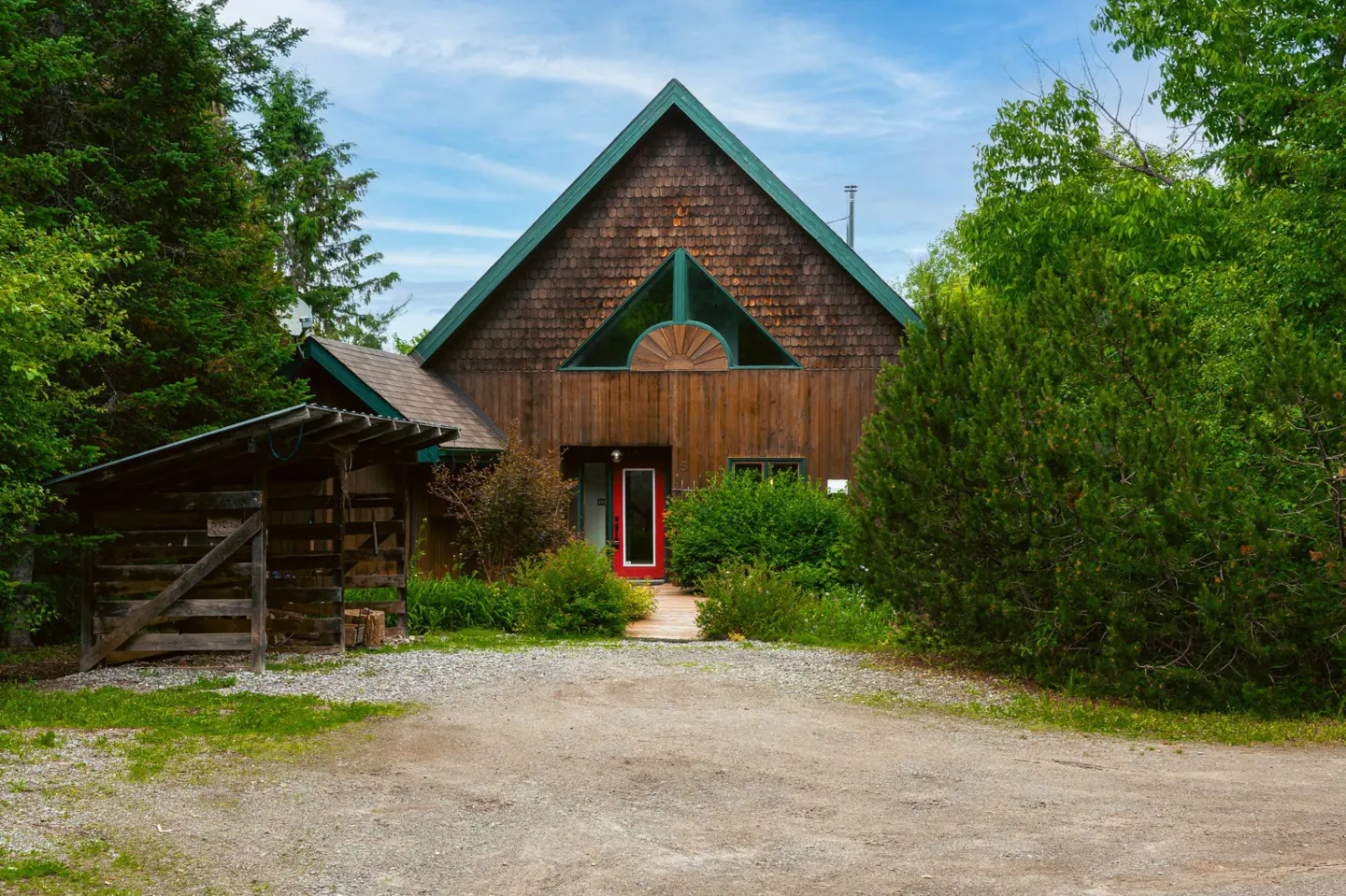 Rustic wooden house with a green-trimmed roof and a bright red door, surrounded by lush greenery and a gravel driveway.