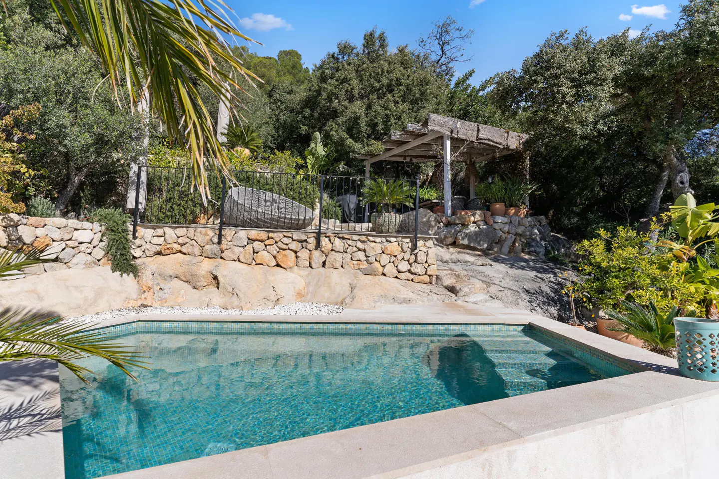 A turquoise tiled pool with stone steps is surrounded by a stone wall, trees, and a pergola.