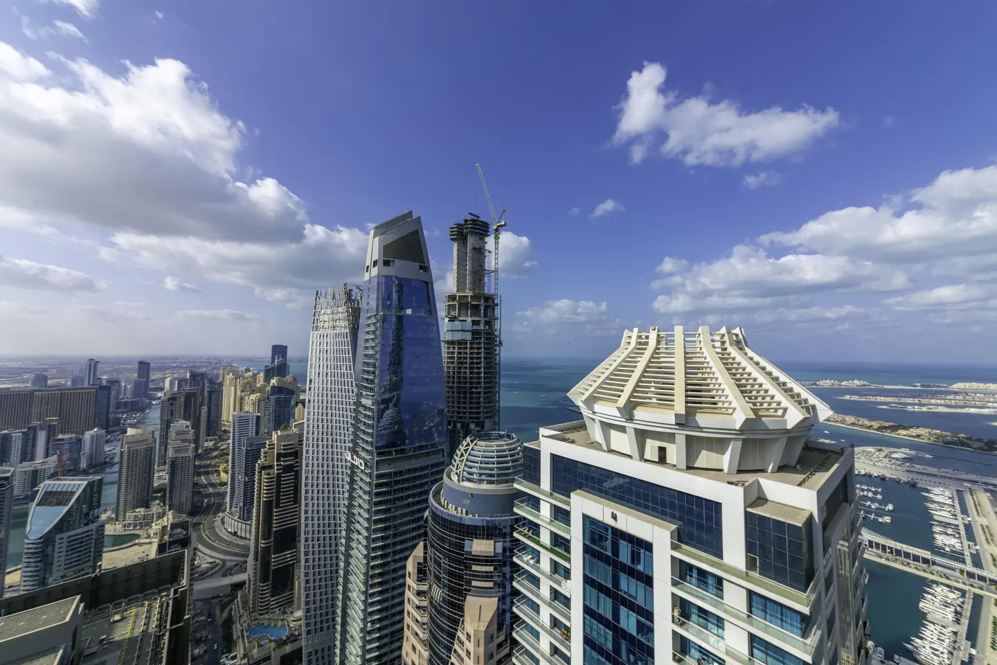 Cityscape view of Dubai with modern skyscrapers, a construction crane, and the sea in the background under a blue sky with white clouds.