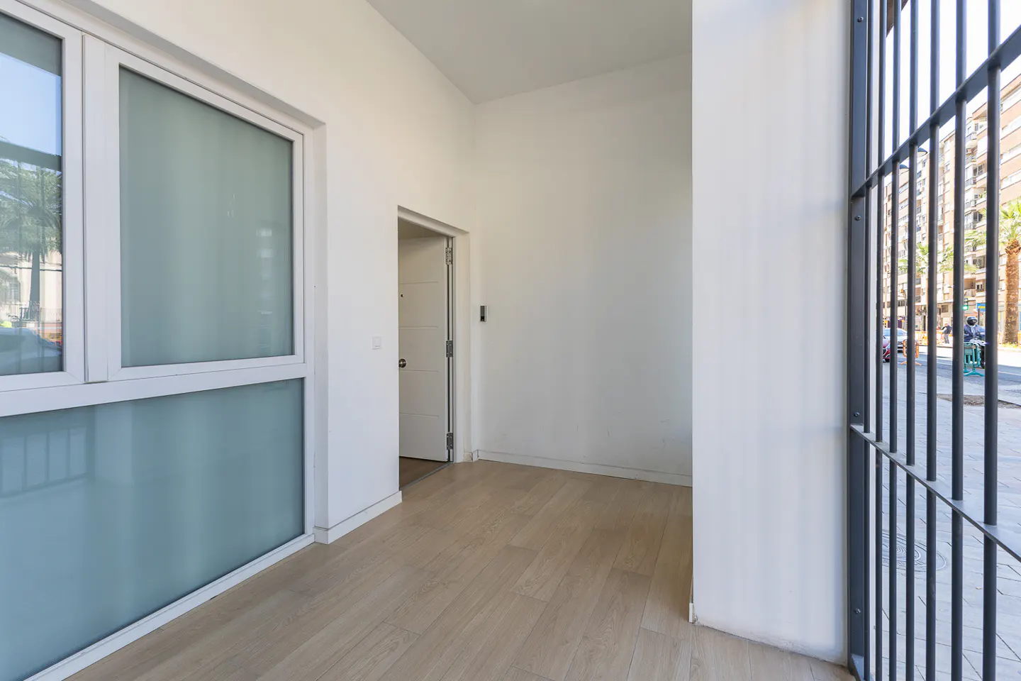 Bright entryway with wood-look floors, white walls, and a large window. A metal gate offers a glimpse of the street outside.
