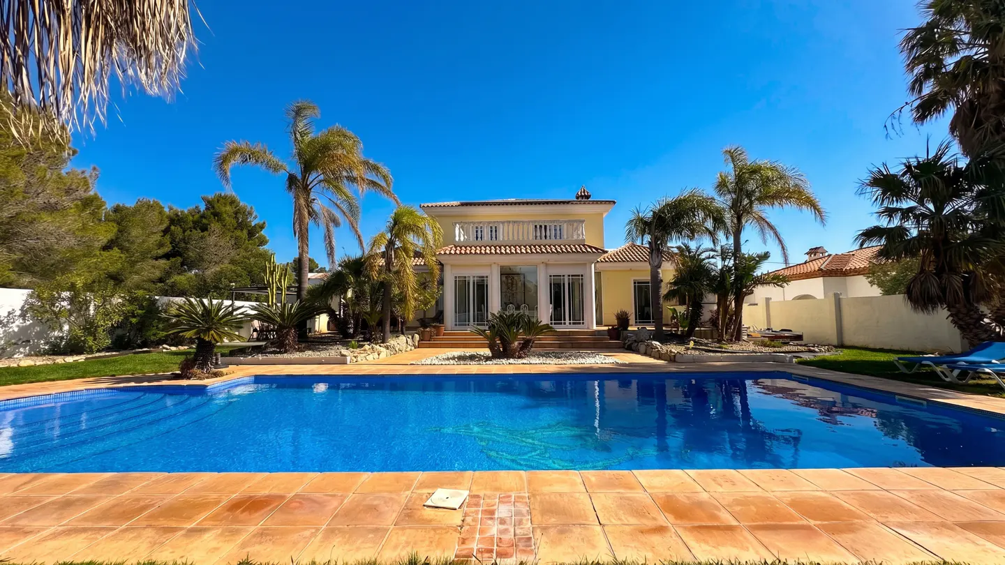 Exterior of a yellow two-story house with a blue swimming pool and palm trees under a clear blue sky.