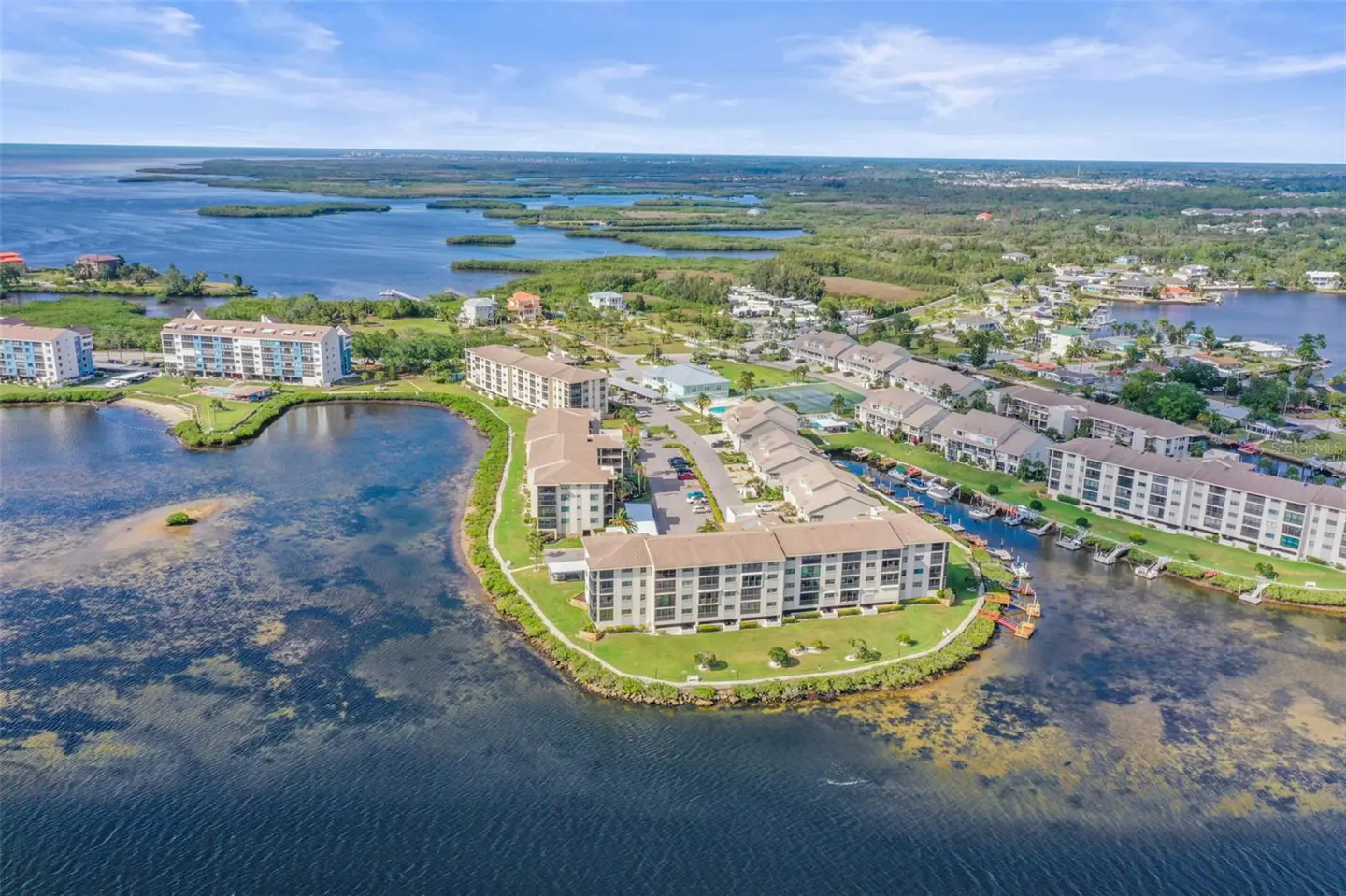 Aerial view of waterfront condos with beige roofs, surrounded by green lawns and blue water. Boats are docked along a canal.