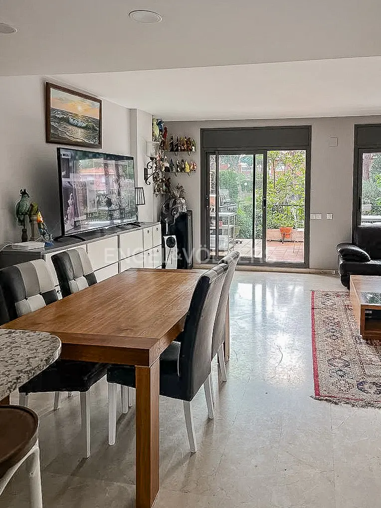 Open-concept living and dining area with a wood table, gray chairs, and a large TV. Sliding glass doors lead to a patio with greenery.