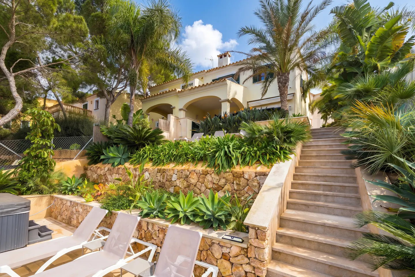 Exterior view of a tan two-story house with a stone retaining wall, palm trees, and lounge chairs.