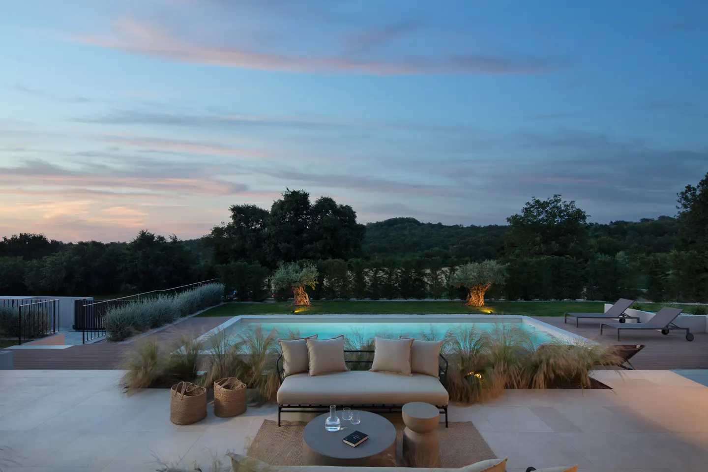 Outdoor patio with a beige couch, table, and pool at dusk. Trees and a colorful sky are in the background.