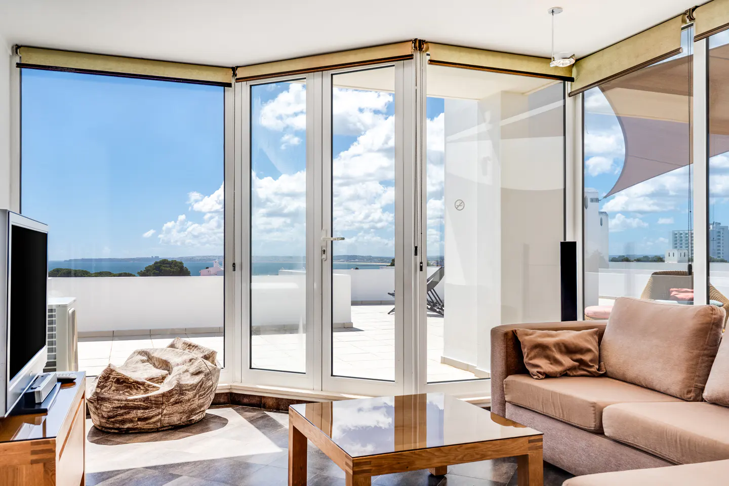 Bright living room with a beige sofa, wooden table, and large windows showing a blue sky and ocean view.