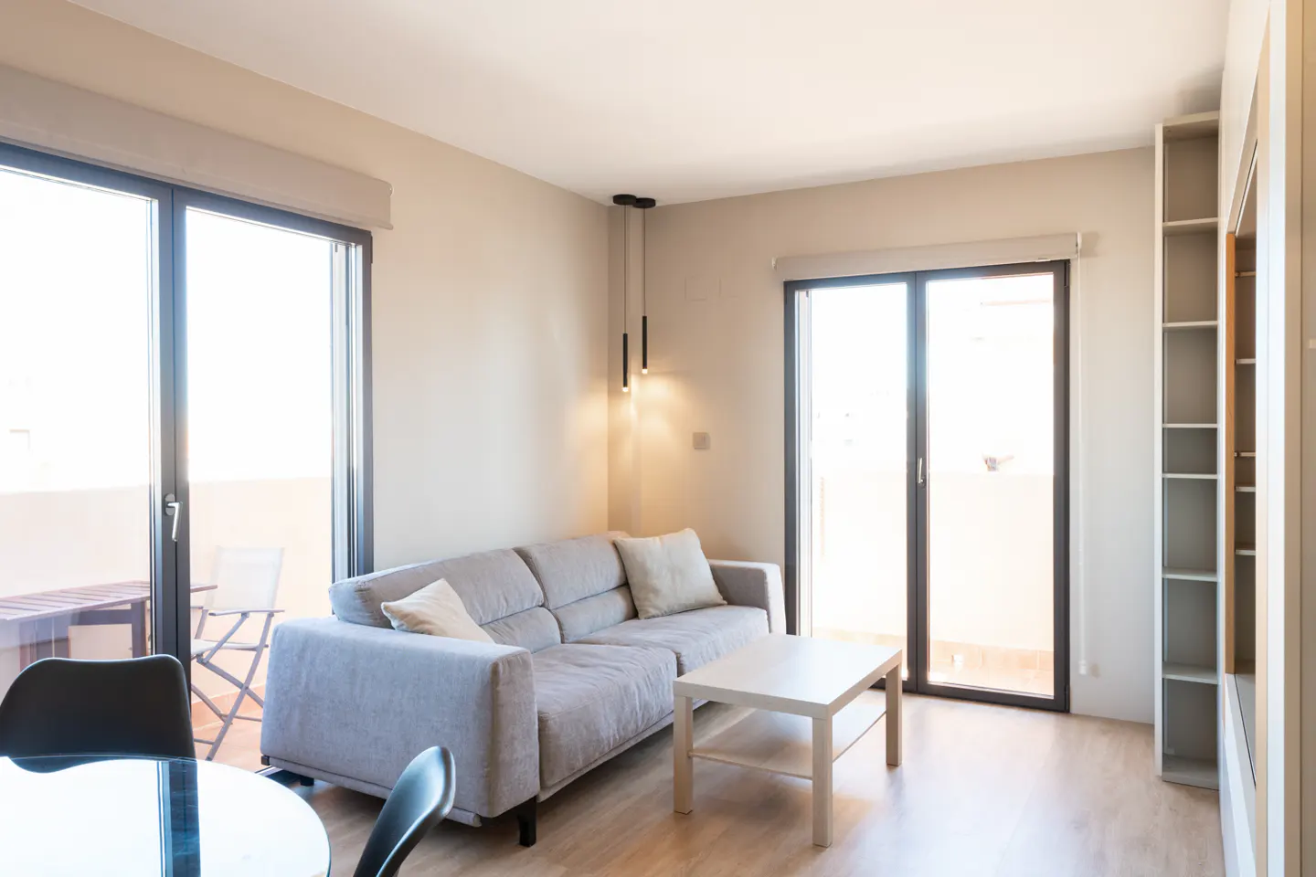 Bright living room with a gray sofa, white coffee table, and floor-to-ceiling windows leading to a balcony. A modern light fixture hangs above.