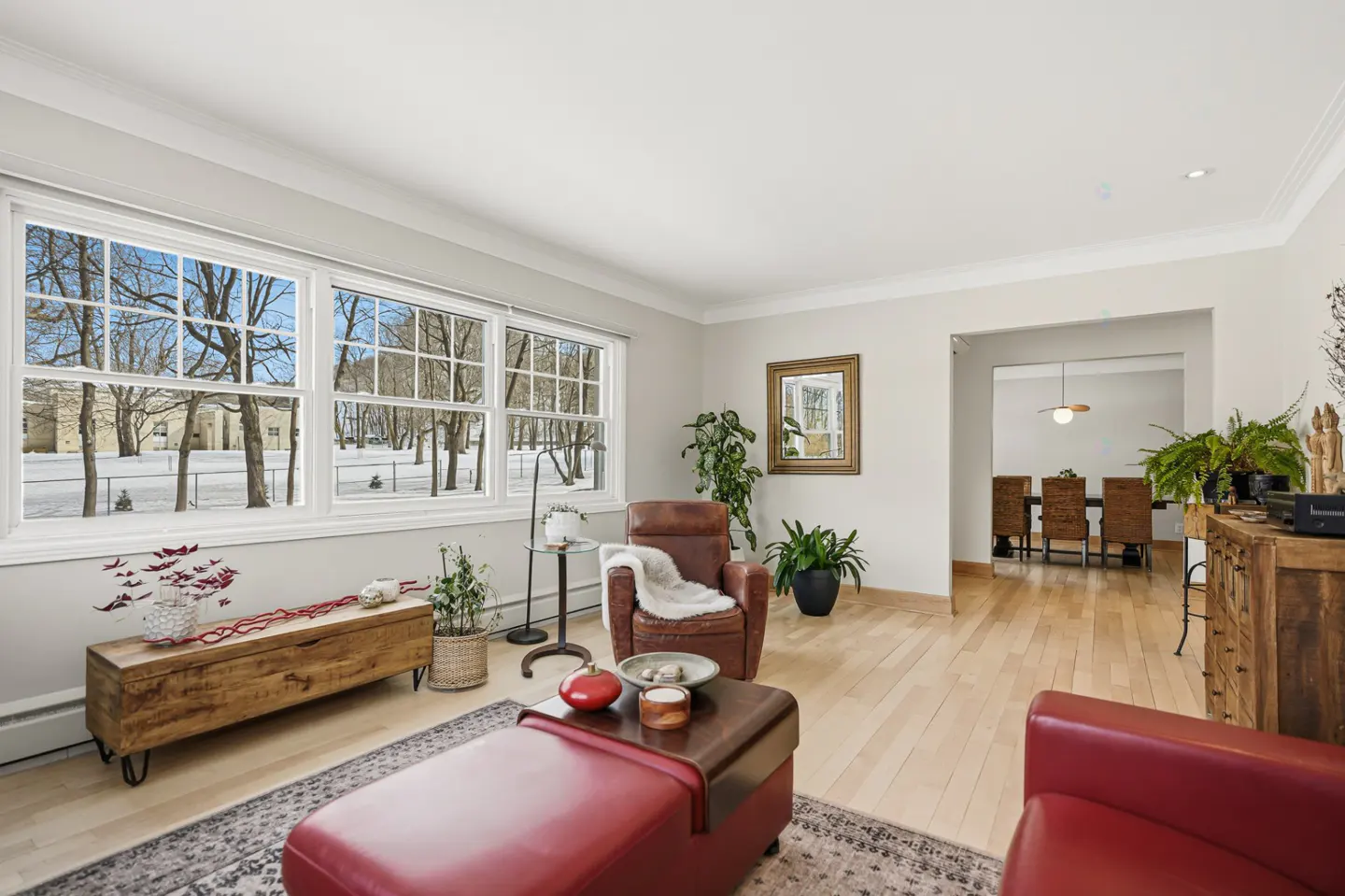 Bright living room with hardwood floors, large windows showing a snowy landscape, and red leather furniture. A dining room is visible in the background.