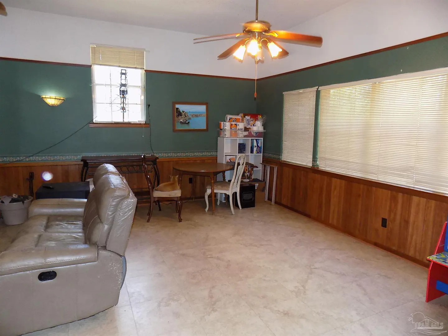A living room with green walls, wood paneling, and a beige recliner. A round table with chairs sits near a white shelf filled with items. A ceiling fan hangs above.