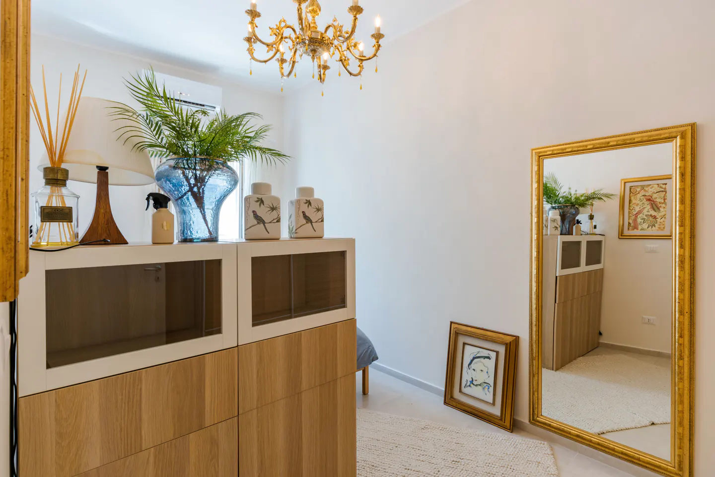 Bedroom interior with a gold chandelier, a white and wood cabinet with decor, and a gold-framed mirror reflecting the room.