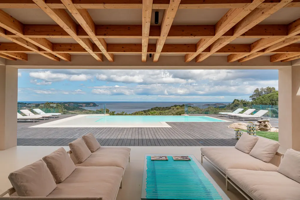 Modern patio with beige sofas and a blue glass coffee table, overlooking a swimming pool and the ocean under a cloudy sky.