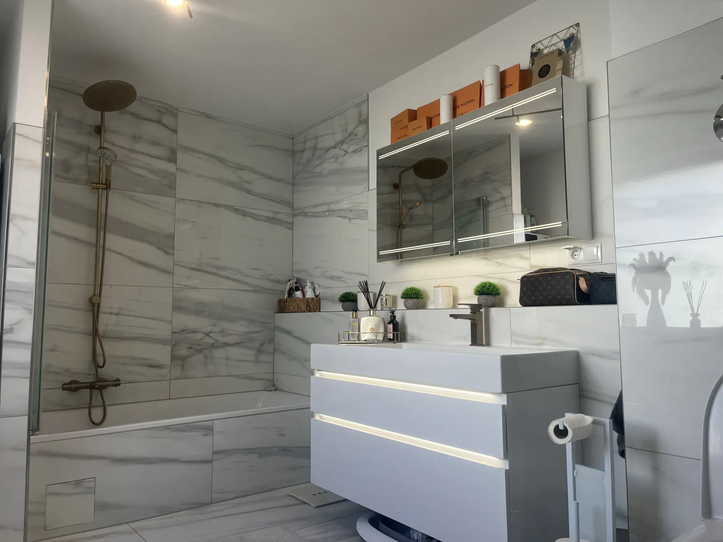 Modern bathroom with marble walls, white vanity with LED lights, and gold shower fixtures.