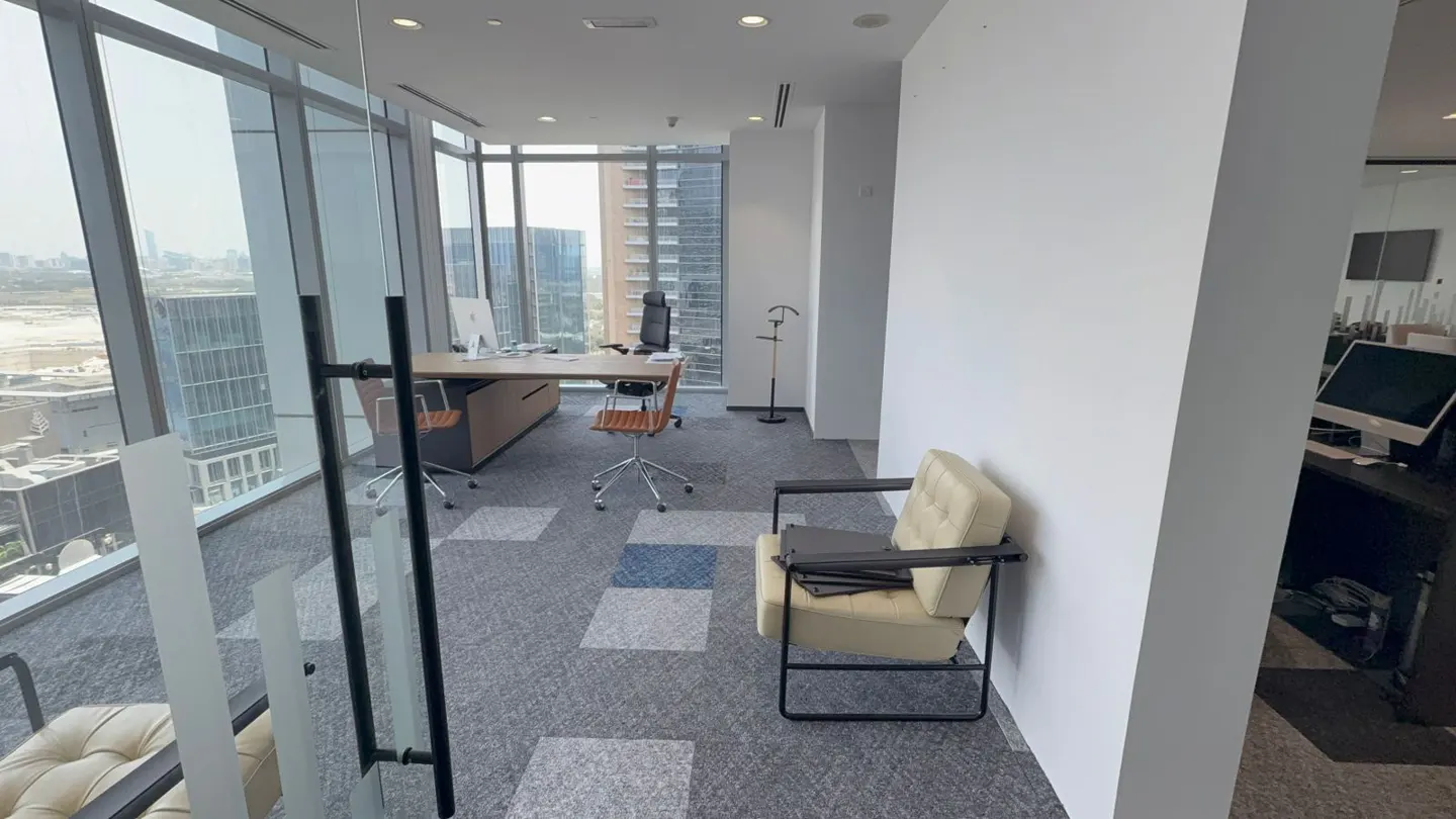 Modern office space with floor-to-ceiling windows, a desk with a computer, and a beige chair on a gray patterned carpet.