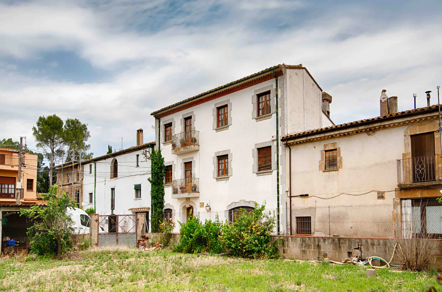 A three-story white building with balconies and brown shutters stands next to older stone houses under a cloudy sky.