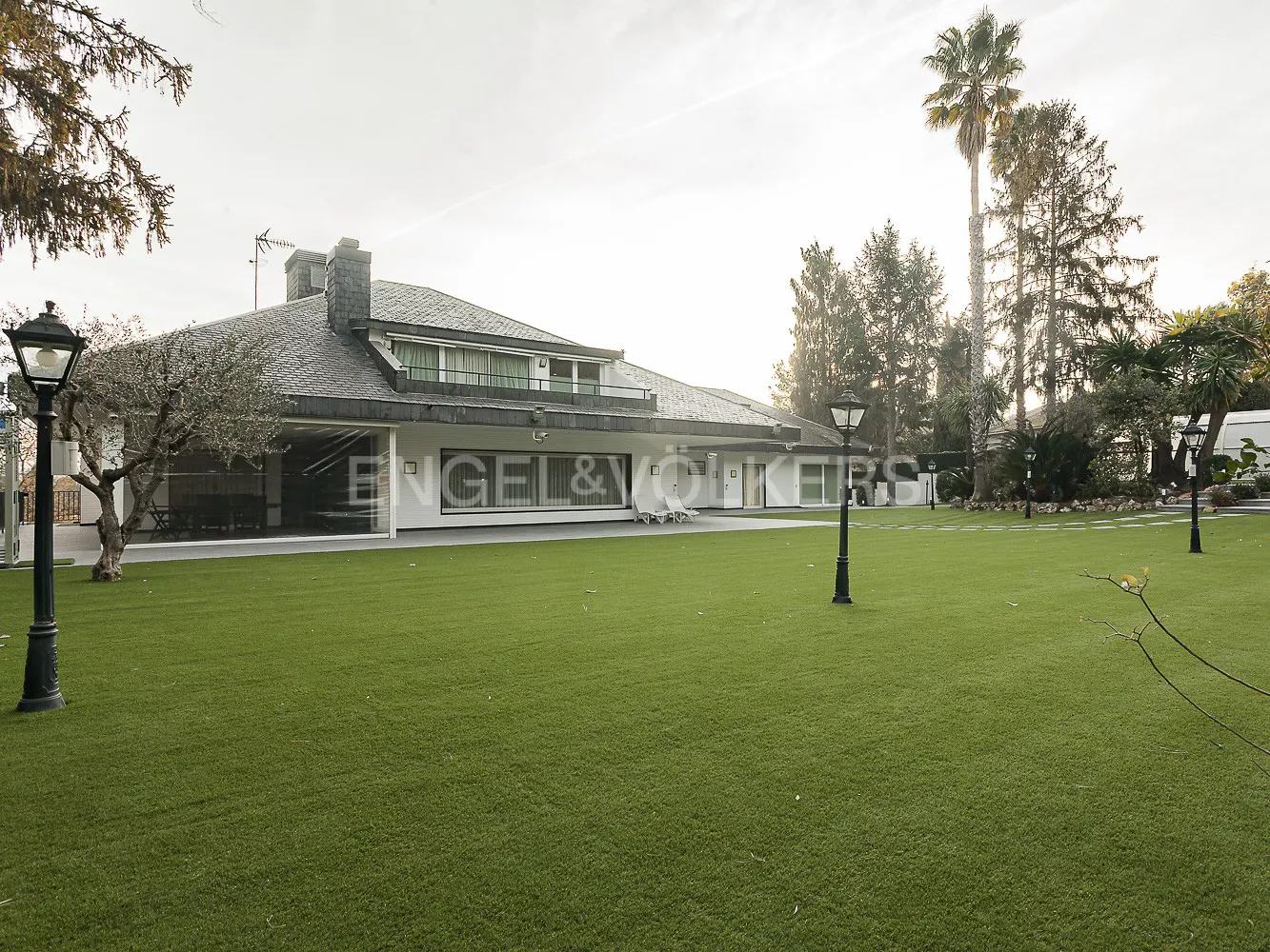 Exterior view of a white, single-story house with a gray roof and a large green lawn. Palm trees and lampposts are visible.