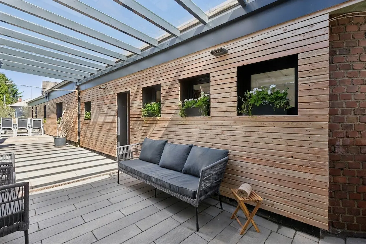 Outdoor patio with gray wicker sofa, wooden wall with flower boxes, and a pergola above.