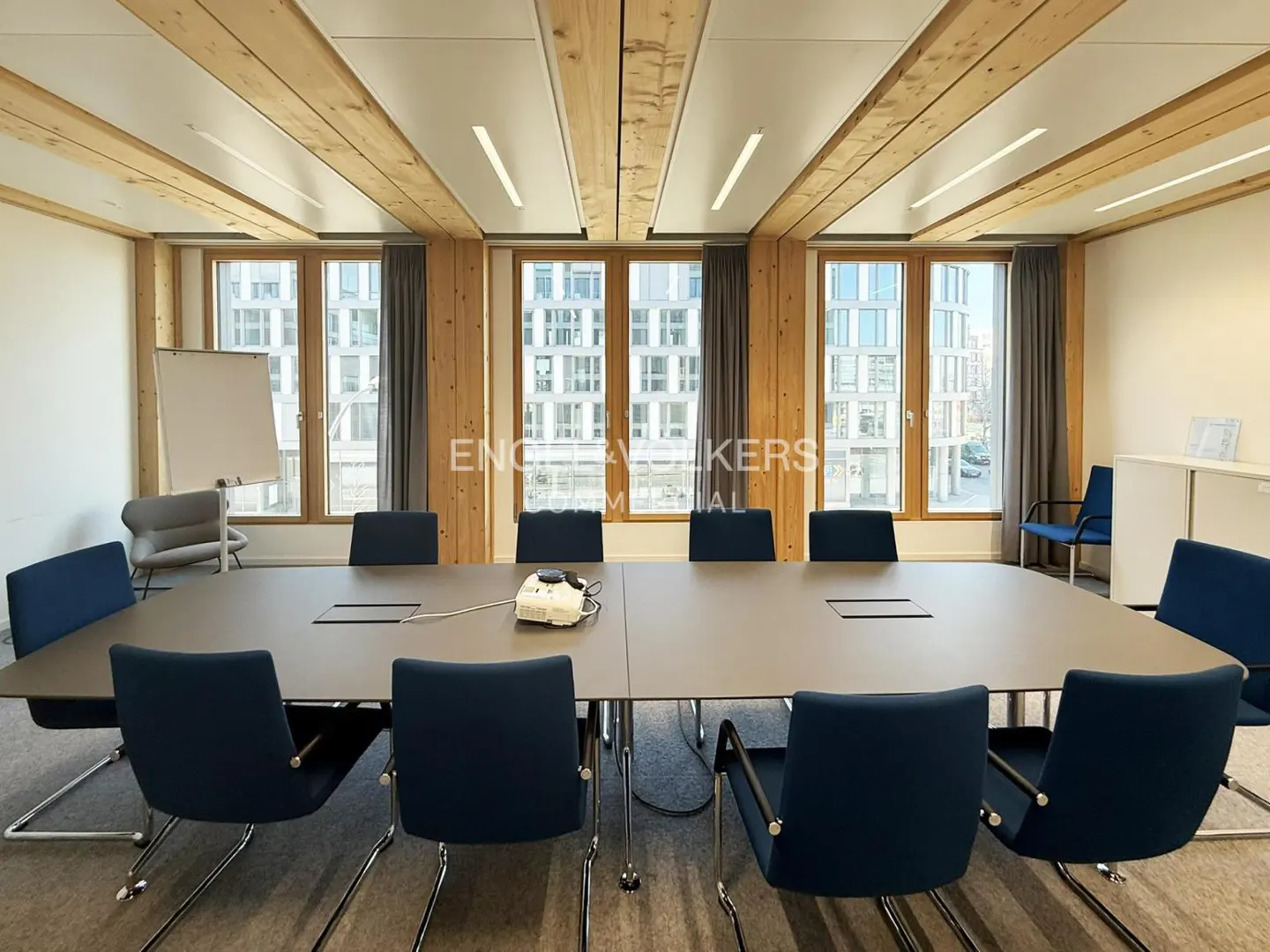 Conference room with a large gray table, blue chairs, and wooden beams on the ceiling. Windows overlook a city.