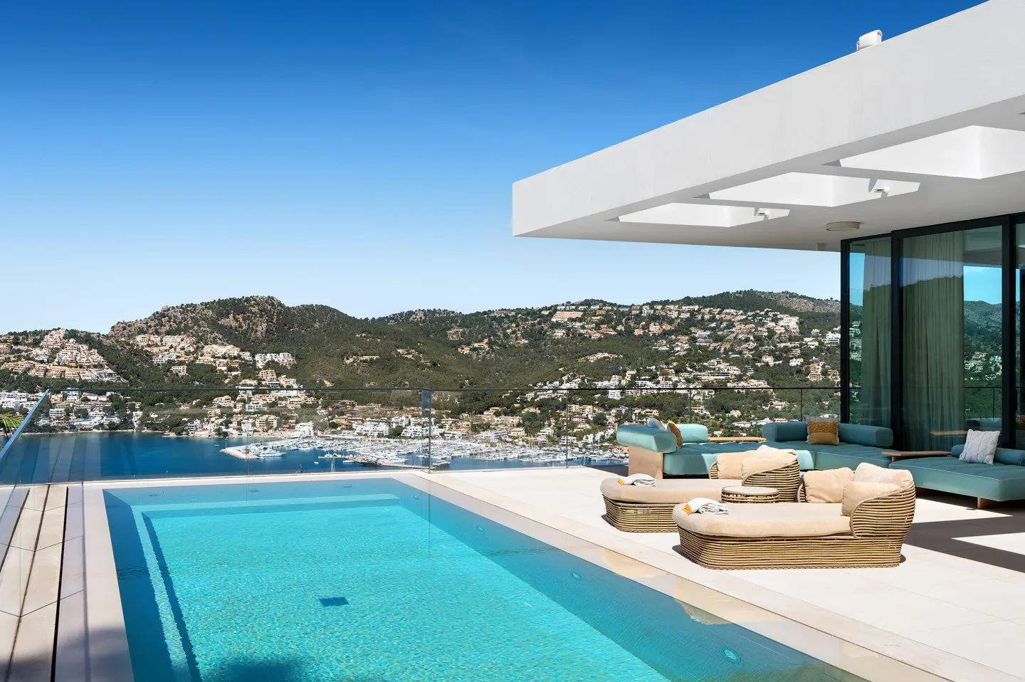 Luxury villa with a pool overlooking a bay. Lounge chairs and sofas sit on the patio. Mountains and a clear blue sky are in the background.