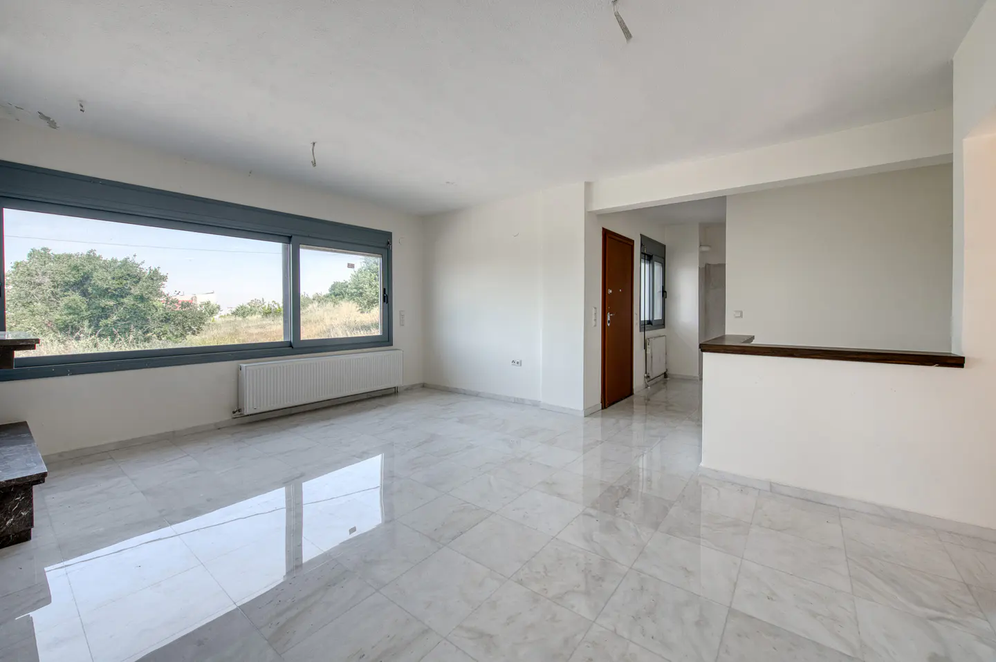 Empty room with white marble floors, large windows, a radiator, and a brown door leading to a hallway.