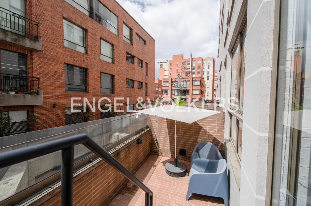 A balcony with a blue chair and white umbrella, surrounded by brick buildings under a cloudy sky. Engel & Volkers logo visible.