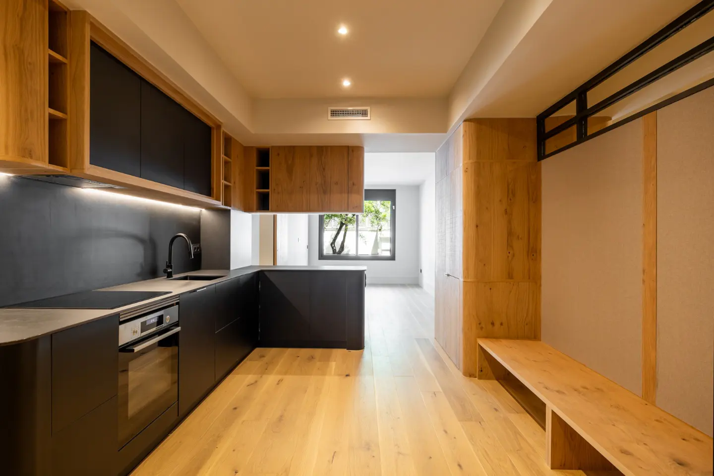 A modern kitchen with black cabinets, wood accents, and a light wood floor. A bench is built into the wall on the right. A window is visible in the background.