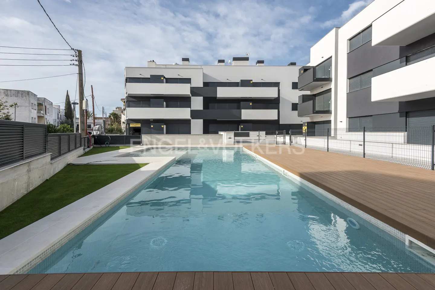 Modern apartment complex with a pool. The building is white and gray, with a wooden deck and green grass surrounding the pool.