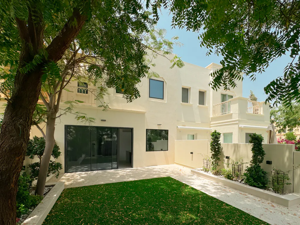 Backyard view of a two-story cream house with a green lawn, trees, and a balcony. Sliding glass doors lead to the interior.