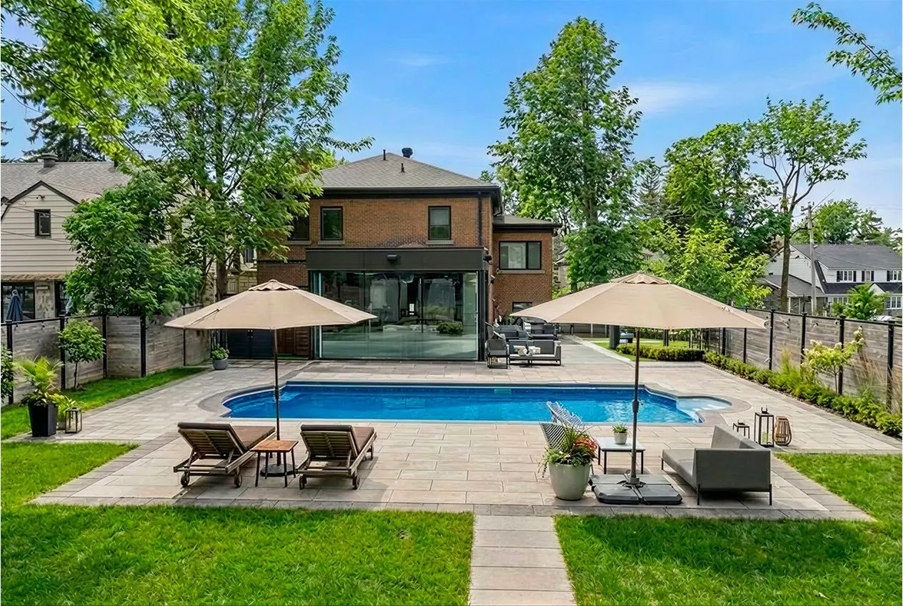 Backyard view of a brick house with a blue pool, lounge chairs, and patio furniture under beige umbrellas.